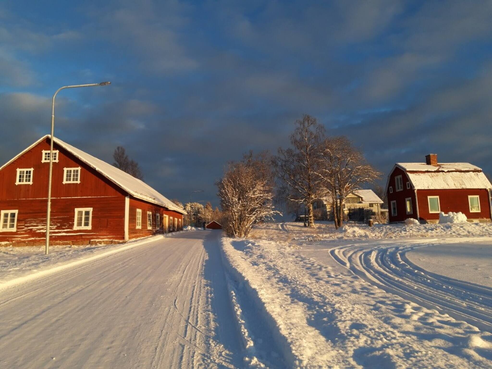 A snow-covered rural road with red wooden buildings, including a barn and a house, under a cloudy sky.