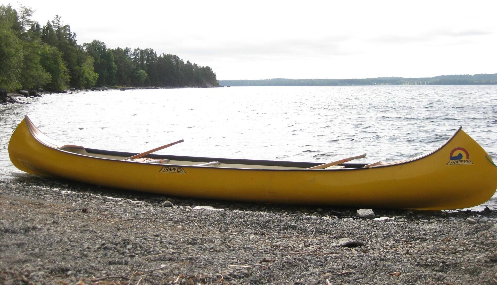 A yellow canoe with a logo on the side, resting on a pebble beach by a calm body of water.