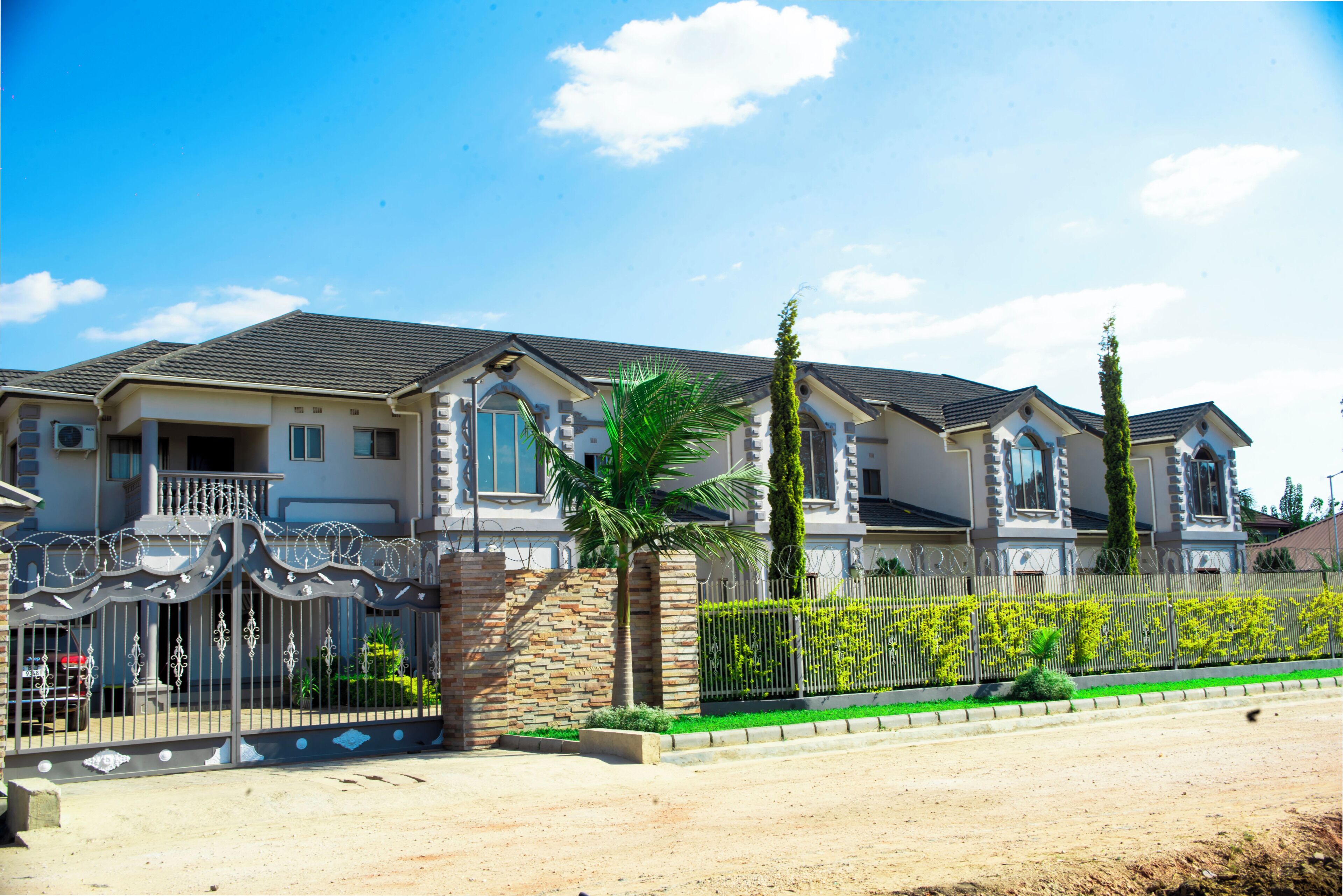 A multi-story residential building with a gated entrance, a stone wall, and a well-maintained lawn with a palm tree.