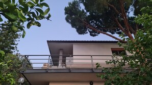 A modern two-story building with a stone facade, a balcony with a metal railing, and a large tree in the background.