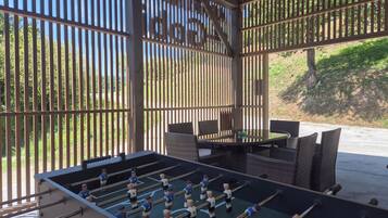 Espaço ao ar livre com área de estar, mesa de futebol de mesa, móveis de vime e vista através de uma cerca de madeira.