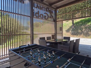 Un área de estar al aire libre con mesa de fútbolín, mobiliario de mimbre y vista a través de una cerca de madera.