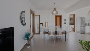 A modern living room with a flat-screen TV, a dining area with white chairs, a kitchen with a refrigerator, and a hallway leading to another room.