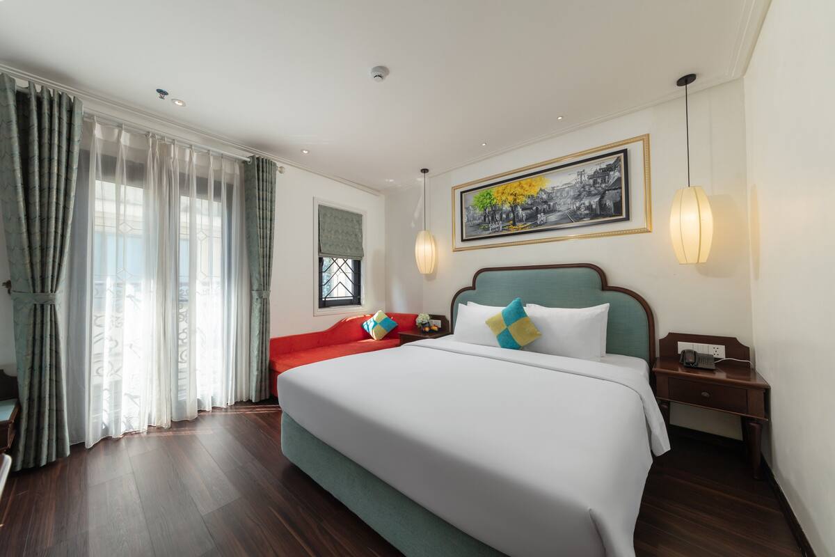 A hotel room with a large bed, a nightstand, a red sofa, and a painting on the wall.