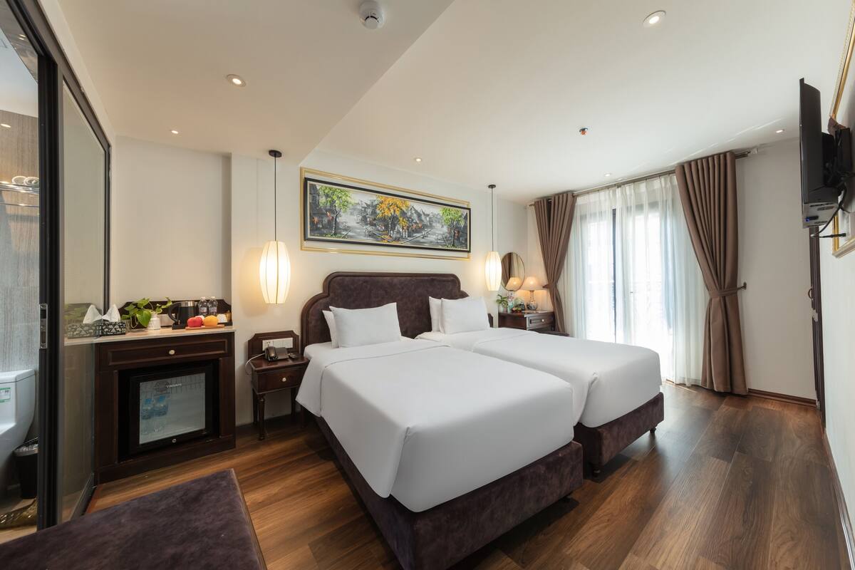 A hotel room with two beds, a TV, a desk with a computer, a fireplace, and a large window with curtains.