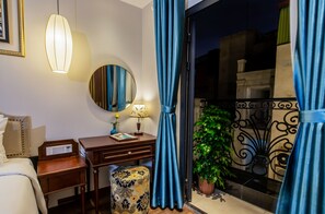 A hotel room with a bed, bedside table, lamp, mirror, and a view of the outside balcony.