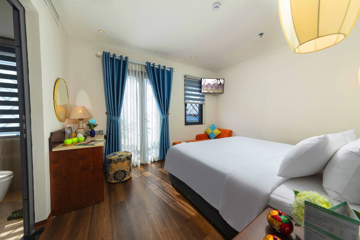 A hotel room with a bed, a desk with a lamp, a chair, a small table with fruit, and a television on the wall.
