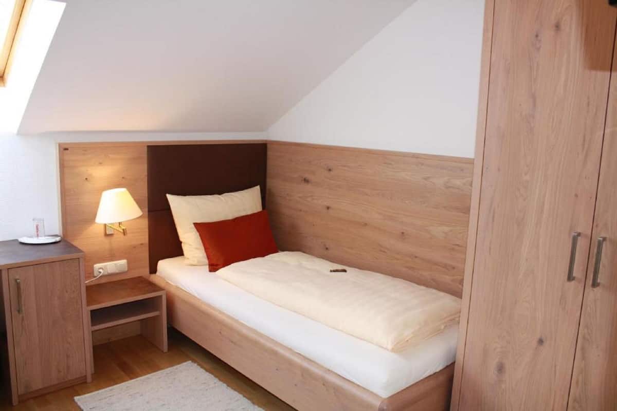 A small, single-bed room with a sloped ceiling, a wooden headboard, a bedside table with a lamp, and a wardrobe.