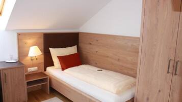 A small, single-bed room with a sloped ceiling, a wooden headboard, a bedside table with a lamp, and a wardrobe.