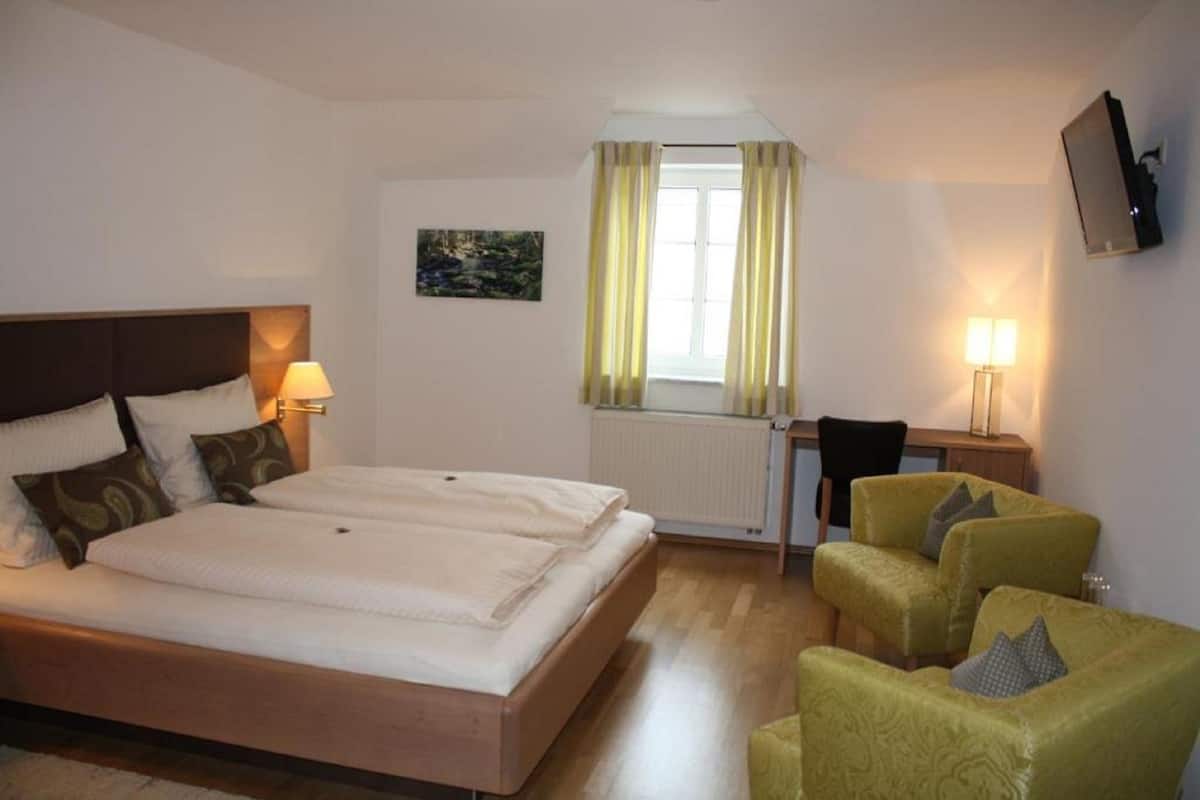 A hotel room with a double bed, a desk, two chairs, a TV, and a window with curtains.
