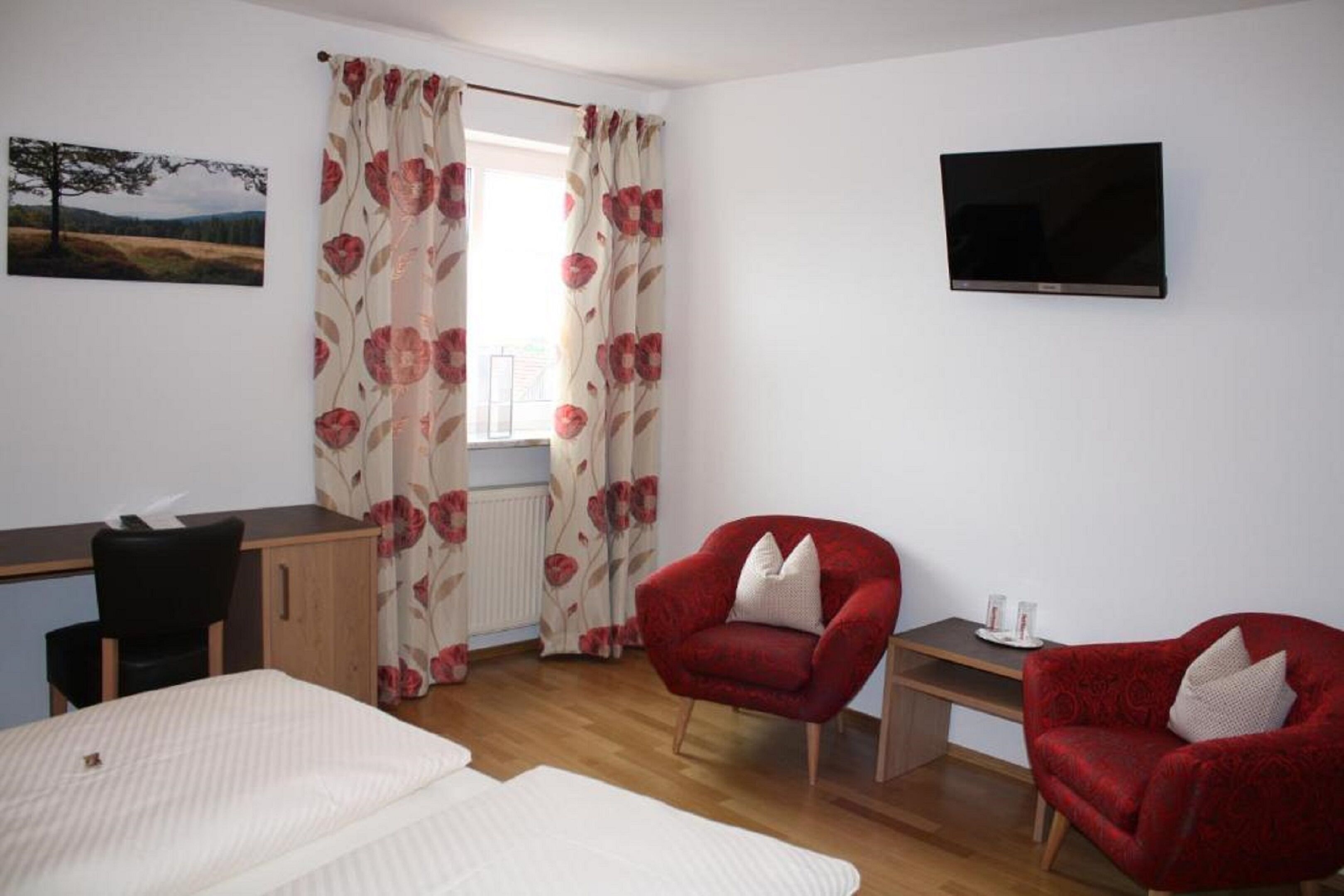 A bedroom with a bed, a desk, two red armchairs, a television, and a window with floral curtains.