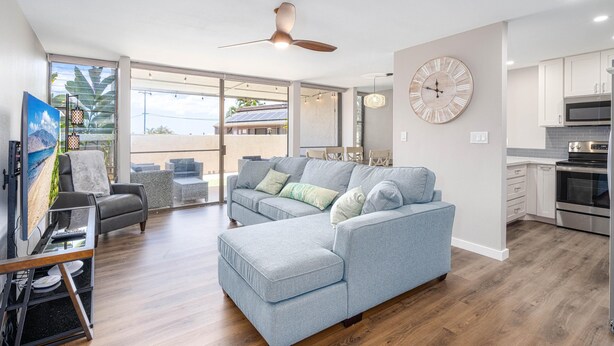 A modern living room with a grey sofa, a dining area with a table and chairs, a kitchen with white cabinets, and a large clock on the wall. - Newly Remodeled Steps to Sugar Beach! (Kihei)