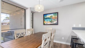 A dining area with a wooden table and white chairs, a painting of a beach scene, and a hanging light fixture. - Newly Remodeled Steps to Sugar Beach! (Kihei)