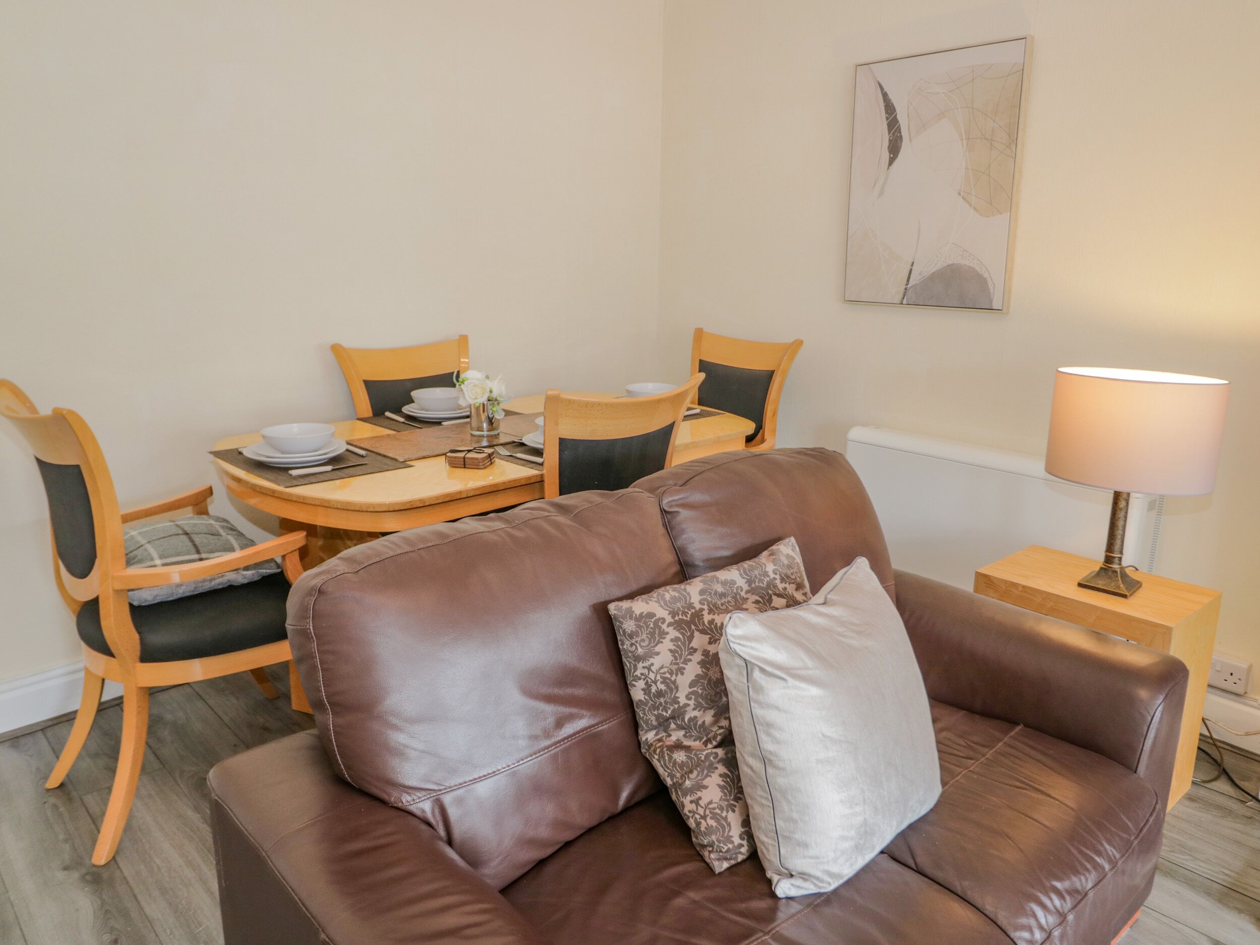 A brown leather sofa with cushions, a wooden dining table with chairs, a lamp, and a painting on the wall.