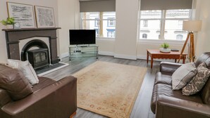 A living room with a fireplace, a television on a glass stand, a sofa, a coffee table, and large windows with a view of buildings. - Gerrard House Apartment (Llandudno)
