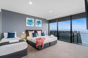 Two beds with white and blue bedding, a large window offering a panoramic ocean view, and abstract art pieces adorn this serene hotel room.