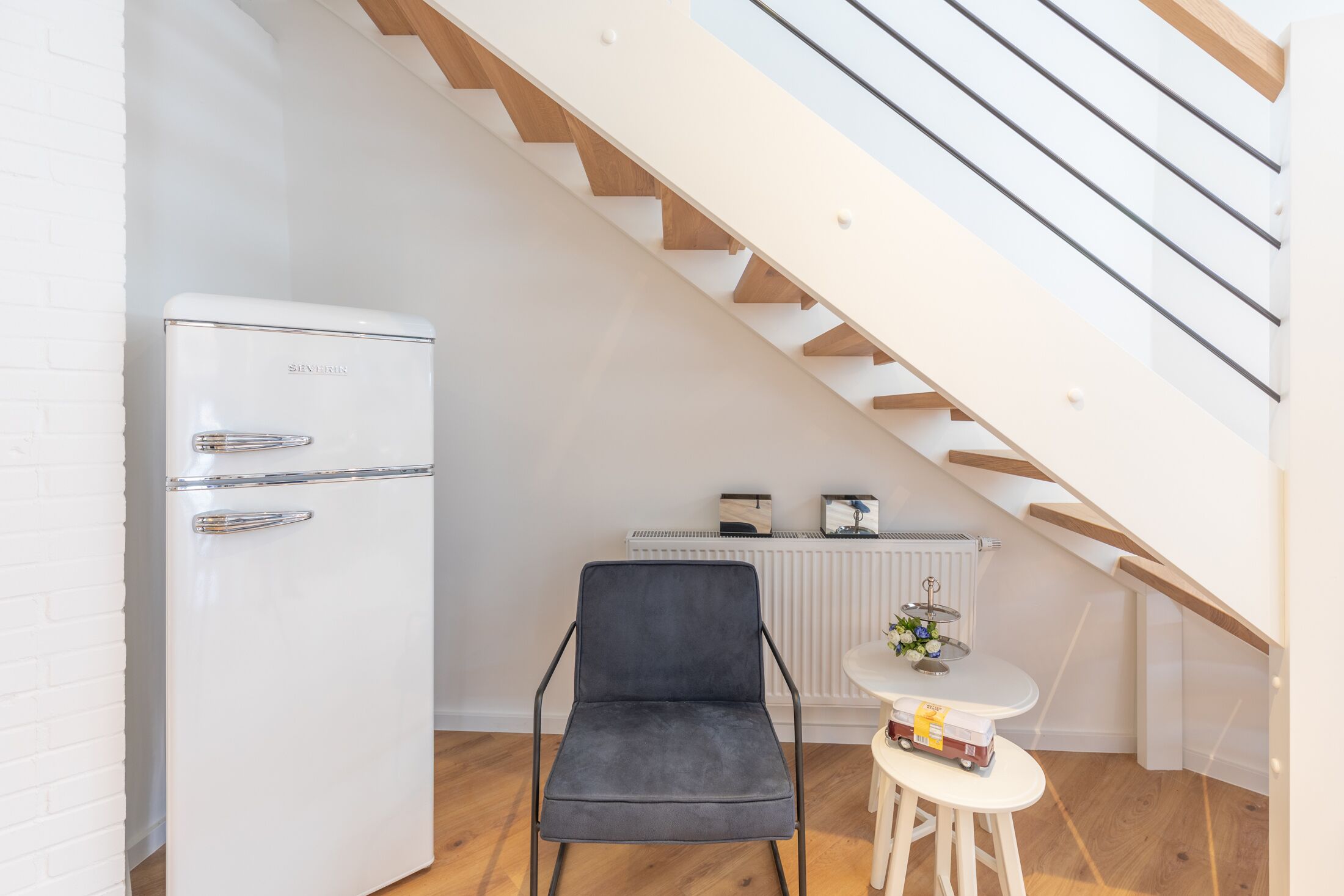 A compact living space with a white refrigerator, a dark grey chair, a small round table with a white base, and a white radiator.