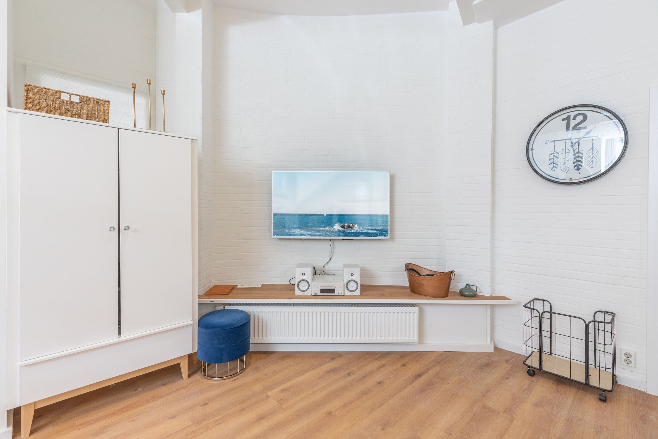 A modern living room with a white brick wall, a mounted television, a round clock, and a wooden floor.