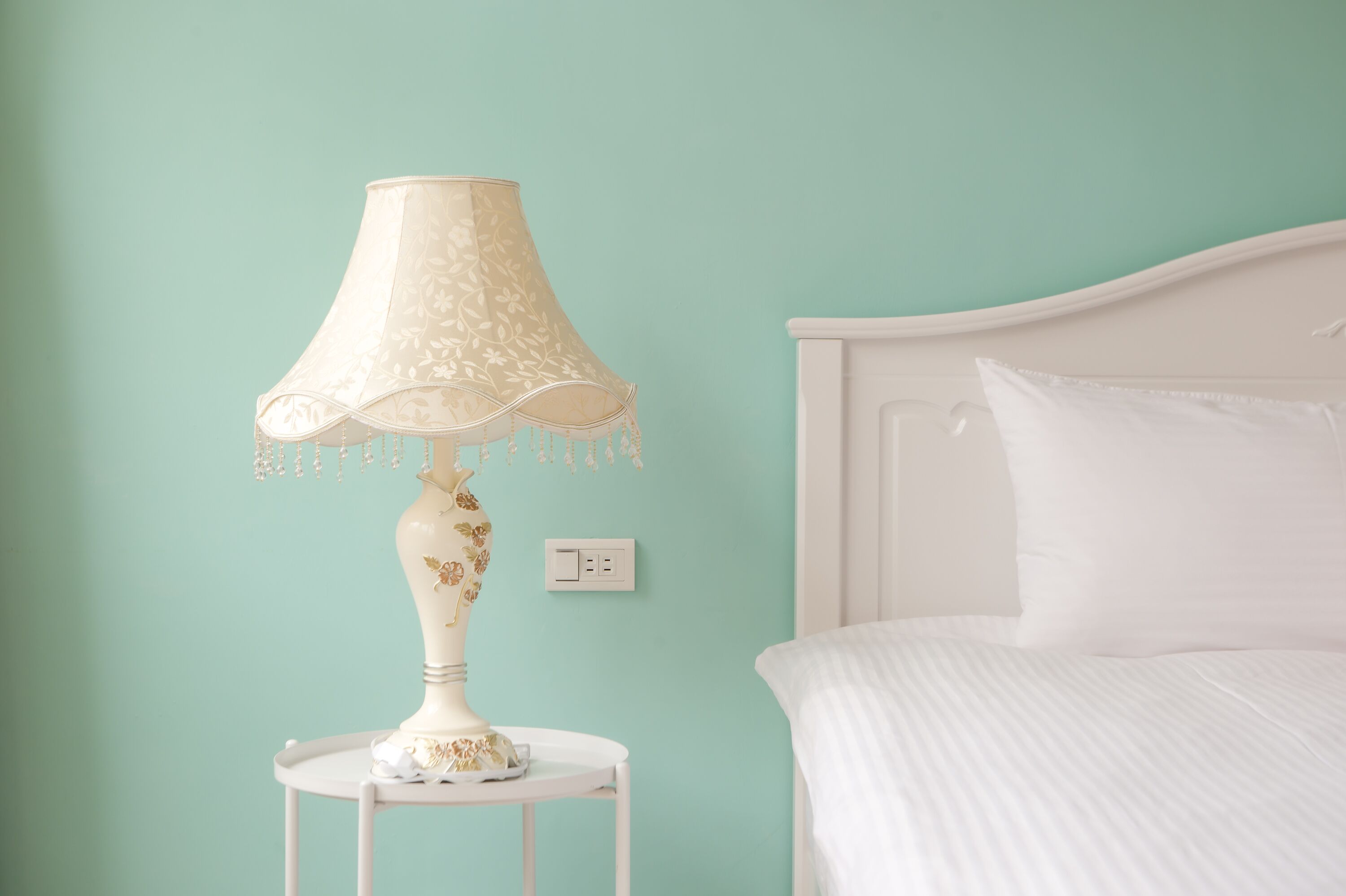 A bed with white bedding, a white nightstand with a lamp, and a light switch on the wall.
