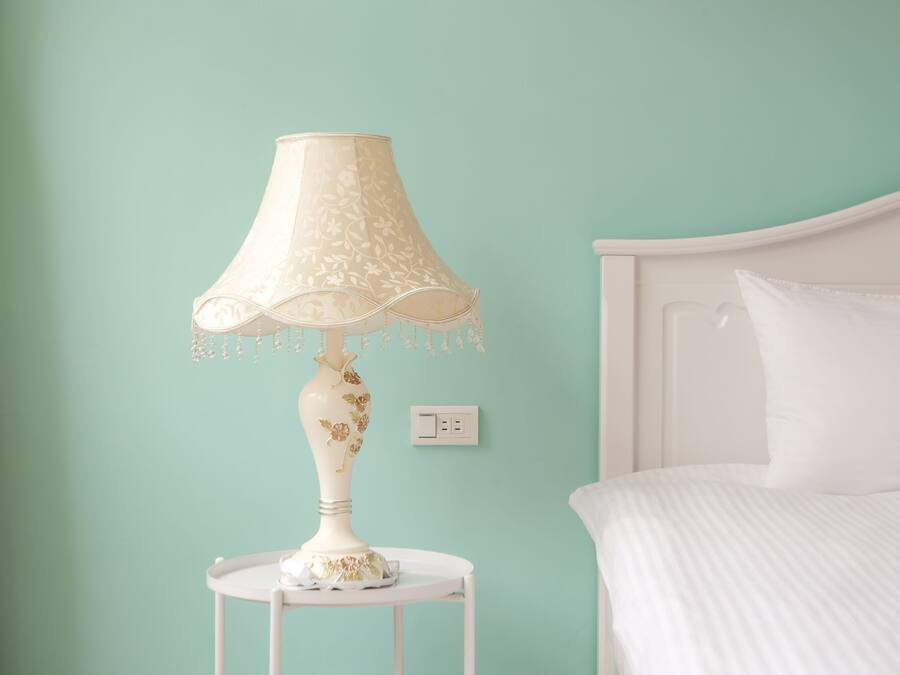 A bed with white bedding, a white nightstand with a lamp, and a light switch on the wall.