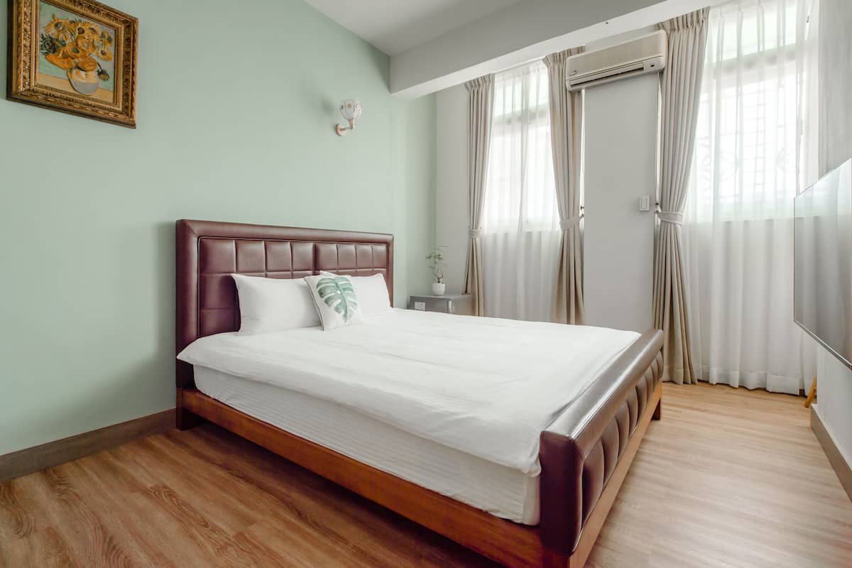A neatly made bed with a wooden headboard, a bedside table with a lamp, and a window with curtains.