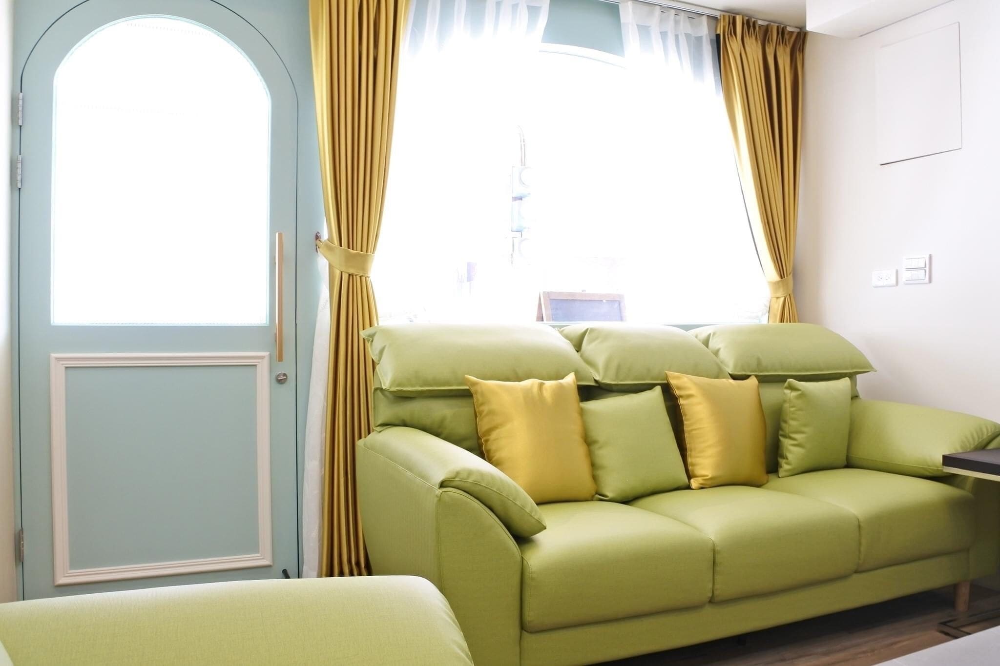 A room with a light blue door, a green sofa with yellow cushions, and yellow curtains.
