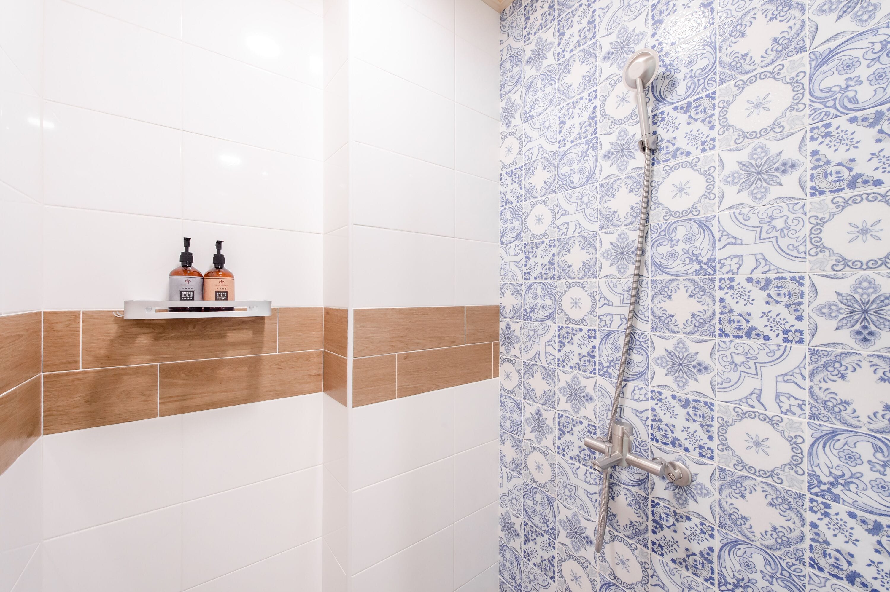 Une salle de bain avec des carreaux blancs et bruns, une douche avec un mur orné d’un motif bleu, et deux bouteilles de shampoing posées sur une étagère.