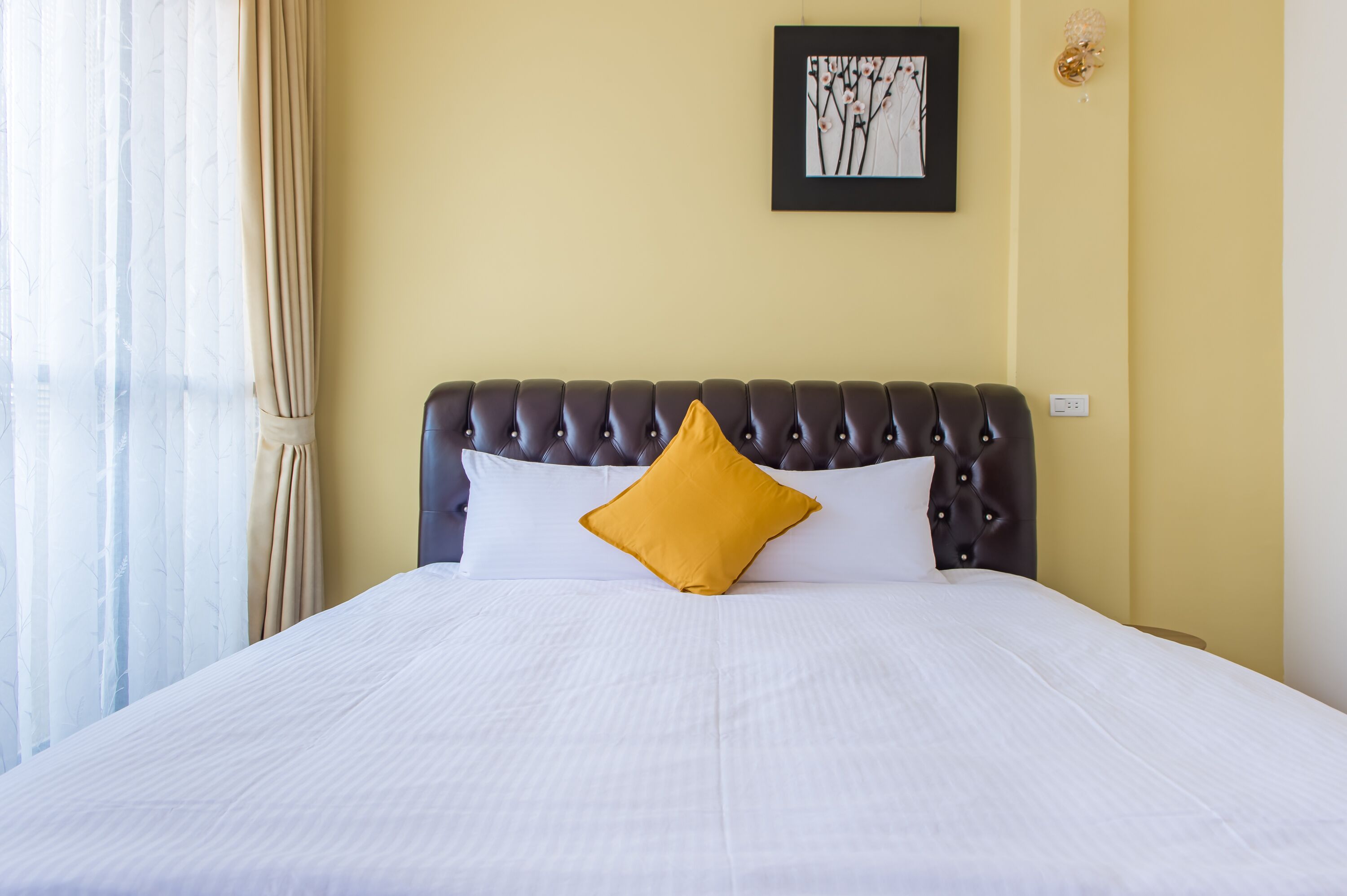 A neatly made bed with white bedding and a single yellow pillow, a dark upholstered headboard, and a framed artwork on the wall.