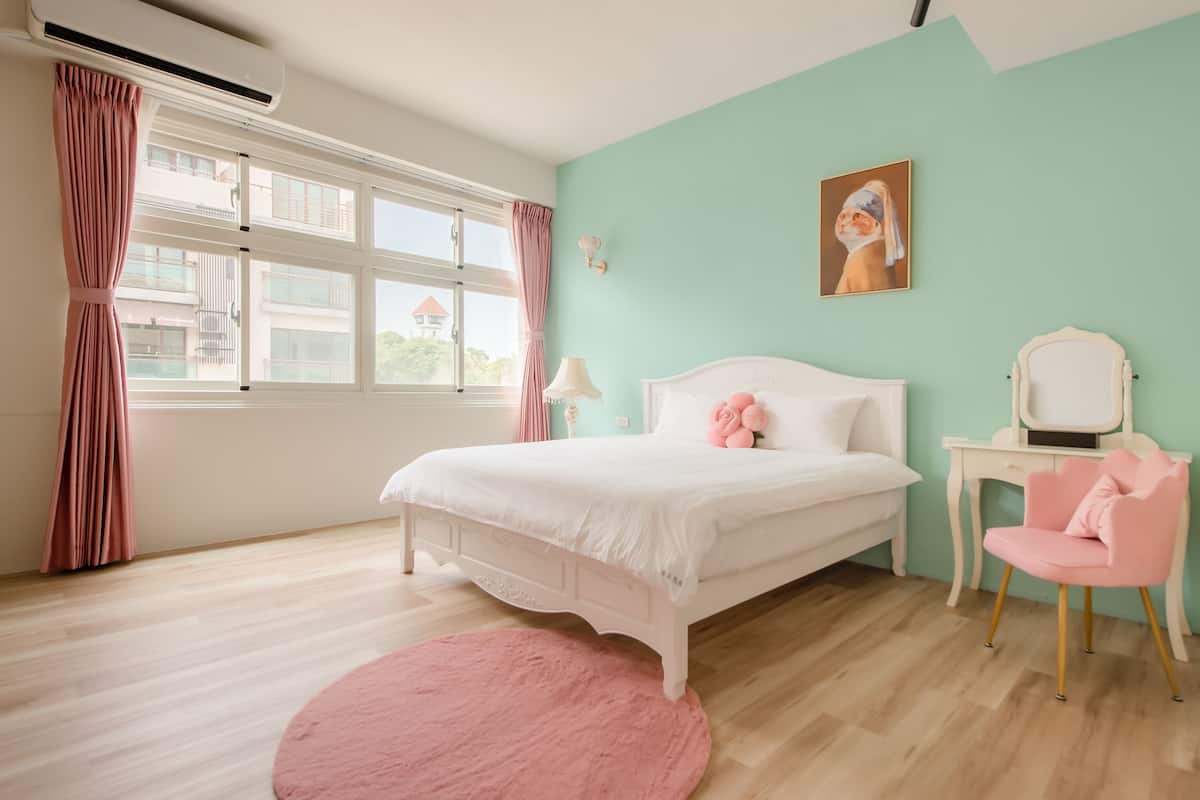 A bedroom with a bed, a desk, a chair, a window with curtains, and a painting on the wall.
