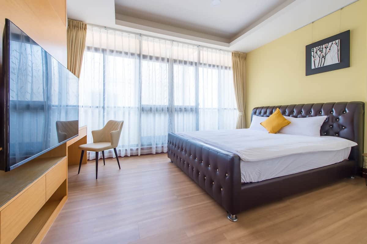 A modern hotel room with a large bed, a flat-screen TV, a desk, and a large window with sheer curtains.