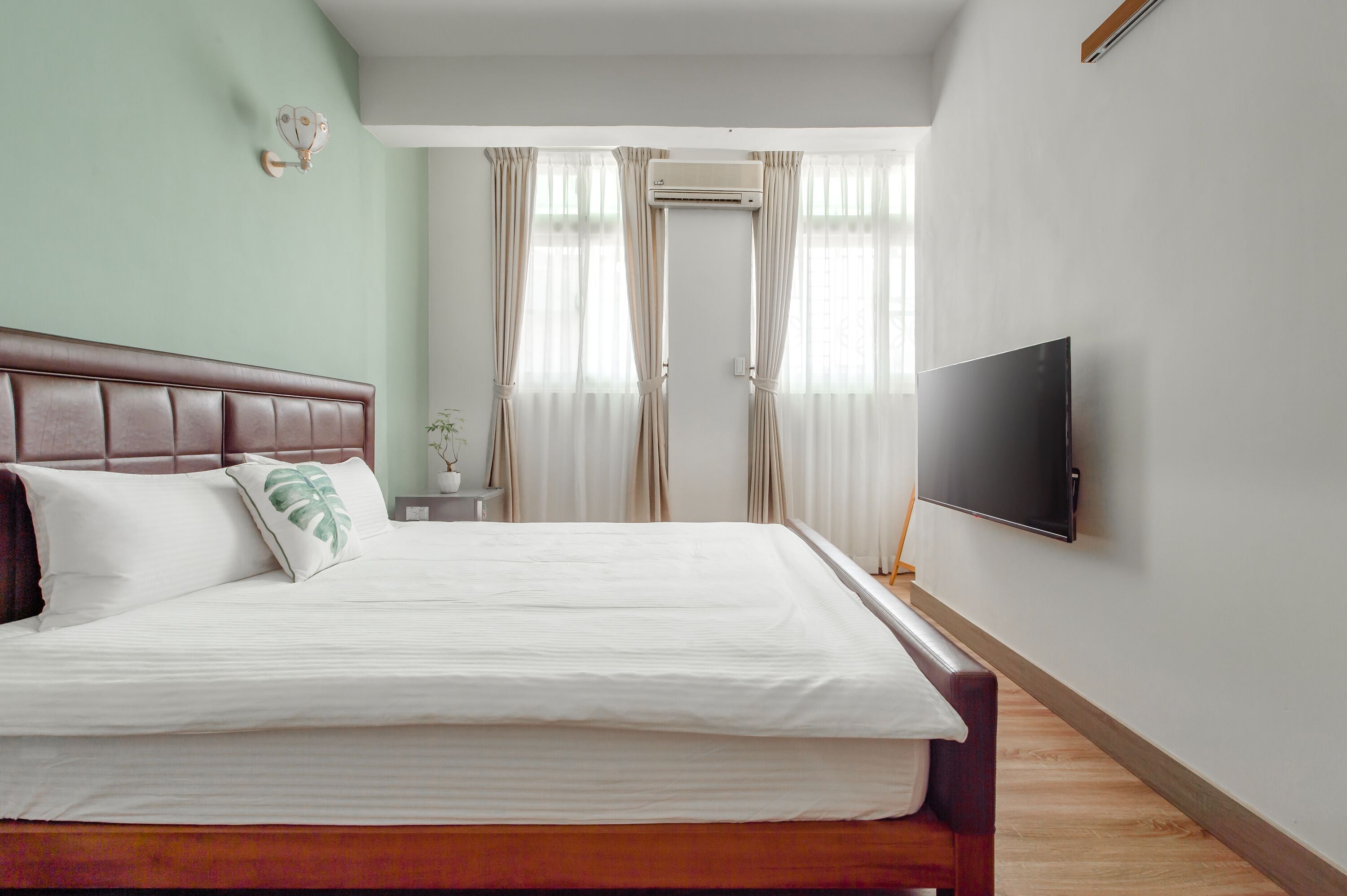A neatly made bed with white linens and a brown headboard, a flat-screen TV mounted on the wall, and a window with sheer curtains.
