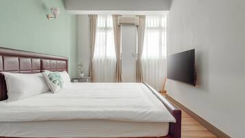 A neatly made bed with white linens and a brown headboard, a flat-screen TV mounted on the wall, and a window with sheer curtains.