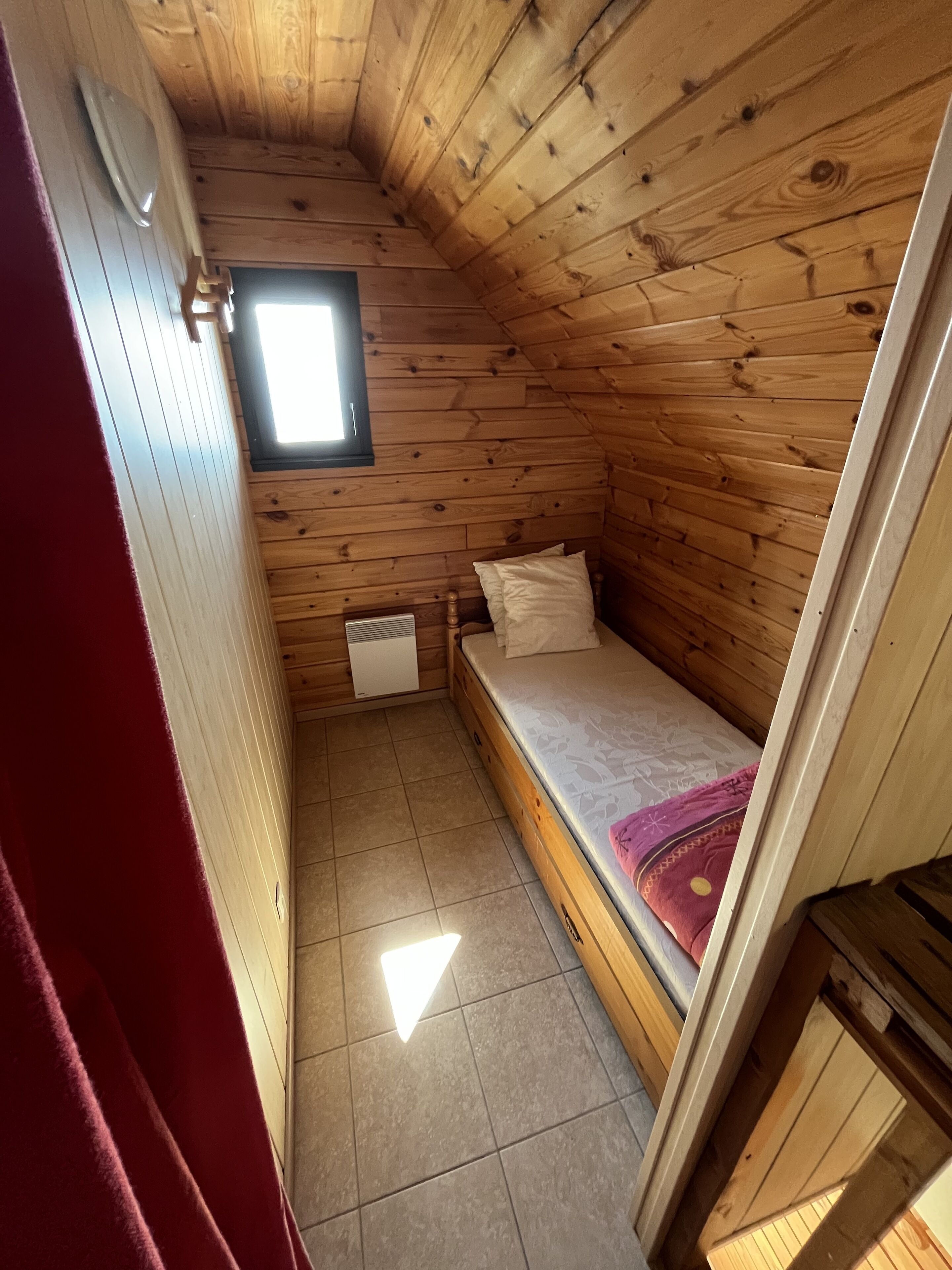 A small, wooden-paneled room with a single bed, a window, and a radiator.