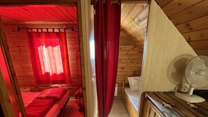 A wooden cabin room with a bed, a small table, and a window with red curtains.