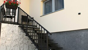 Un escalier avec une rampe noire menant à un palier, une fenêtre avec un rideau et un jardinière avec des fleurs rouges sur la gauche.