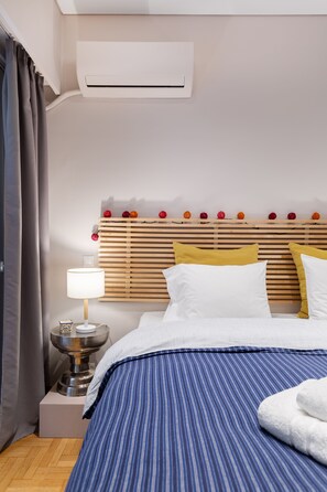 A neatly made bed with a striped blue bedspread, a white pillow, and a yellow pillow. There is a bedside table with a lamp and a decorative item. - Bang in Kolonaki by Athenian Homes (Athens)