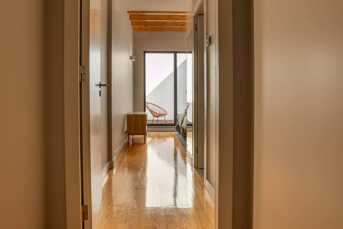 A hallway with wooden flooring, a door leading to a room with a chair and a table, and a view of a balcony.