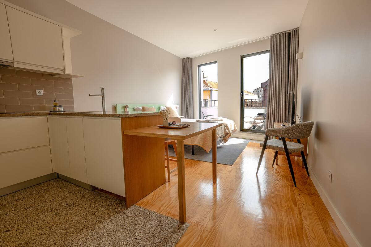 A modern kitchen with a wooden table and chairs, a small dining area, and a view of the outside through a large window.