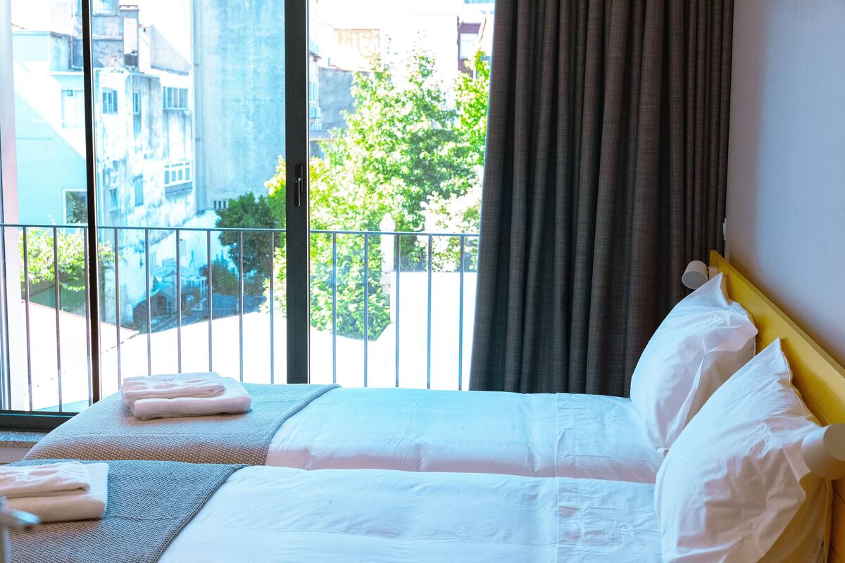 A hotel room with two beds, a balcony, and a view of buildings and trees.