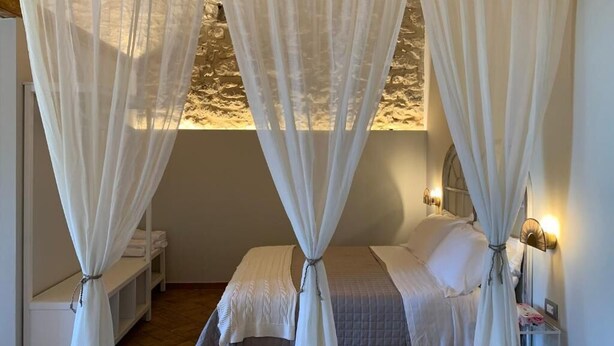 A bedroom with a bed, white curtains, a nightstand, and a wall-mounted lamp. - Masseria Santacatrini - Loft Nocellara (Melilli)