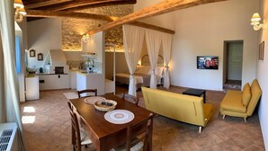 A cozy living area with a wooden dining table, yellow sofa, and a bed with white curtains. - Masseria Santacatrini - Loft Nocellara (Melilli)