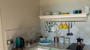 Una cocina compacta con armarios blancos, un hervidor verde azulado, una cafetera y un colgador de ollas con tazas amarillas.