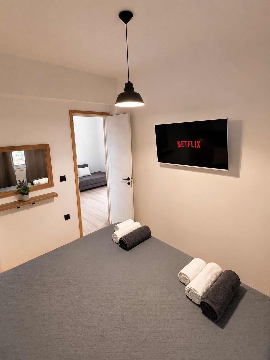 Un dormitorio moderno con una cama grande, un televisor montado en la pared, una mesita de noche con una planta y una puerta que da a otra habitación.