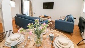 A modern living room with a round dining table set for four, two blue sofas, a flat-screen TV mounted on the wall, and a large window with curtains. - Stabal (Ty Croes)