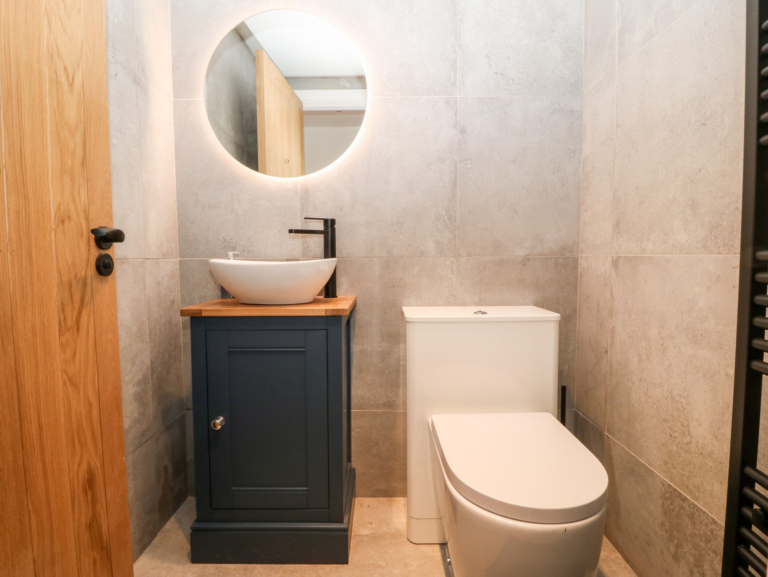 Un baño con un mueble de madera, un lavabo blanco, un grifo negro, un inodoro y un espejo redondo en la pared.