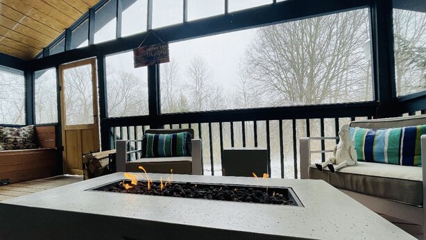 A cozy room with a fireplace, large windows, and a view of trees. - Cozy House with Hot Tub (Macedon)