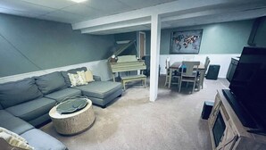 A living room with a grey sofa, a round coffee table, a piano, a dining table with chairs, and a television. - Cozy House with Hot Tub (Macedon)