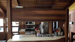 A kitchen with wooden beams, a central island with stools, stainless steel appliances, and a view into the dining area. - Cozy House with Hot Tub (Macedon)