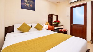 A neatly made bed with white linens and two mustard-colored pillows, a bedside table with a lamp and a vase of red flowers, and a wooden door with a window. - HANZ Boutique Hotel Ben Thanh (Ho Chi Minh City)