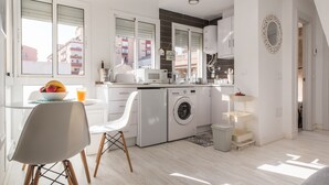 Eine moderne Küche mit Waschmaschine, ein Essbereich mit weißem Stuhl und Blick auf Gebäude durch große Fenster.